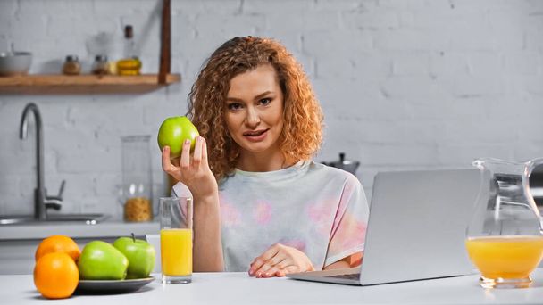 ノートパソコンの近くのカメラとテーブルの上の果物を見ている間にアップルを持っている巻き女性 - 写真・画像