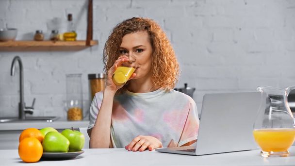 femme bouclée boire du jus d'orange tout en regardant la caméra près de l'ordinateur portable et les fruits sur la table - Photo, image