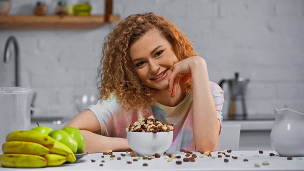 curly woman smiling near pile of corn flakes in bowl  - Fotó, kép