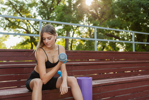 Jeune femme mince en forme faisant de l'exercice haltère tout en étant assis sur tribune en bois à l'extérieur le matin - Photo, image