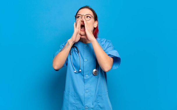 young attractive red hair woman feeling happy, excited and positive, giving a big shout out with hands next to mouth, calling out. hospital nurse concept - 写真・画像