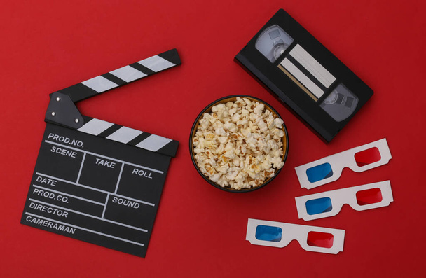 Film tapsoló tábla, popcorn tál, 3D-s szemüveg, videokazetta piros háttérrel. Szórakoztató ipar. Első látásra. Lapos fektetés - Fotó, kép
