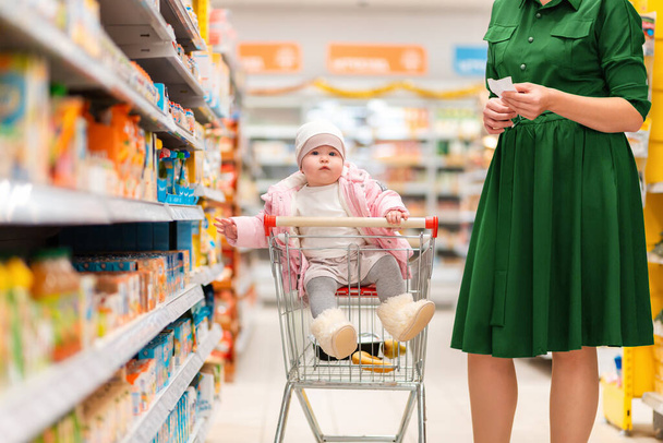 Una madre joven y su bebé sentados en un carrito de comestibles eligen comida en un estante del supermercado. Vista lateral. De cerca. El concepto de compras y crianza. - Foto, Imagen