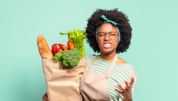 young pretty afro woman looking angry, annoyed and frustrated screaming wtf or whats wrong with you and holding a vegetables bag  - 写真・画像