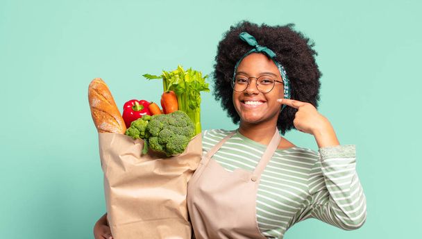 young pretty afro woman making capice or money gesture, telling you to pay your debts! and holding a vegetables bag - Foto, Imagen