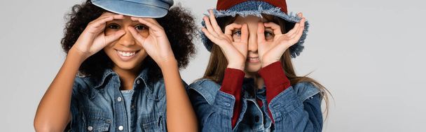 radostné multikulturní děti v džínových šatech a stylových čepicích napodobujících brýle s rukama izolovanýma na šedé, banner - Fotografie, Obrázek