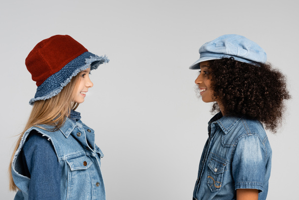 side view of two interracial girls in trendy clothes and hats smiling isolated on grey - Foto, afbeelding