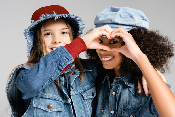 happy multicultural girls in denim clothes and hats showing heart symbol with hands isolated on grey - Foto, Imagem