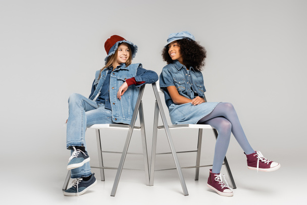 trendy multicultural girls looking at each other while sitting on chairs on grey - Foto, imagen