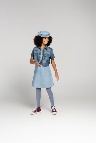 fashionable african american girl in denim clothes, cap and gumshoes posing on grey - Fotó, kép