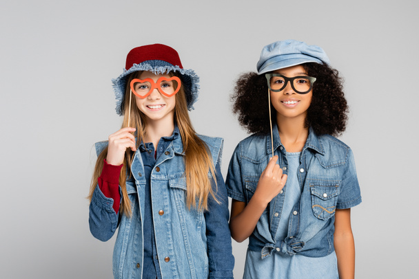 smiling multicultural girls in denim clothes holding party spectacles isolated on grey - Photo, Image