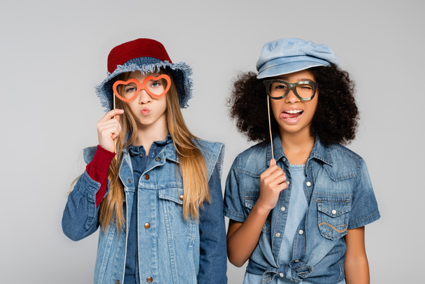 χαρούμενα, κομψά διαφυλετικά κορίτσια που κρατούν γυαλιά κομμένα από χαρτί και γρυλίζουν απομονωμένα σε γκρι - Φωτογραφία, εικόνα