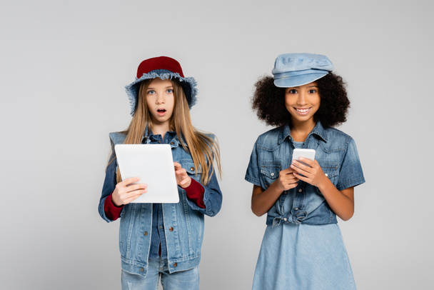izgatott fajok közötti gyerekek divatos ruhák segítségével okostelefon és digitális tabletta elszigetelt szürke - Fotó, kép