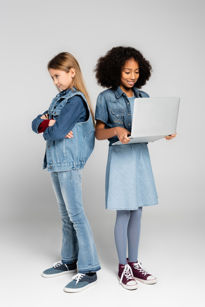 offended girl standing with crossed arms near happy african american friend using laptop on grey - Foto, imagen