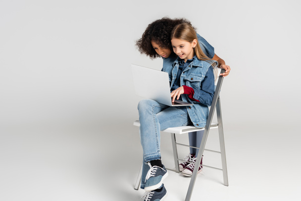 cheerful kid in fashionable denim clothes sitting on chair and using laptop near african american friend on grey - Foto, imagen