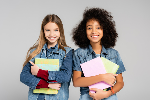 веселые, модные межрасовые школьницы, держащие книги и улыбающиеся в камеру, изолированную на сером - Фото, изображение