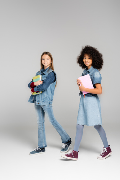trendy interracial schoolgirls in denim clothes and gumshoes walking with textbooks on grey - Photo, Image