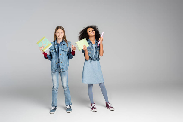 full length view of fashionable multicultural schoolgirls standing with books on grey - Fotografie, Obrázek