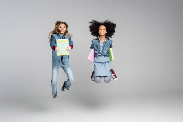 веселые межрасовые школьники в стильной джинсовой одежде прыгают с книгами на сером - Фото, изображение