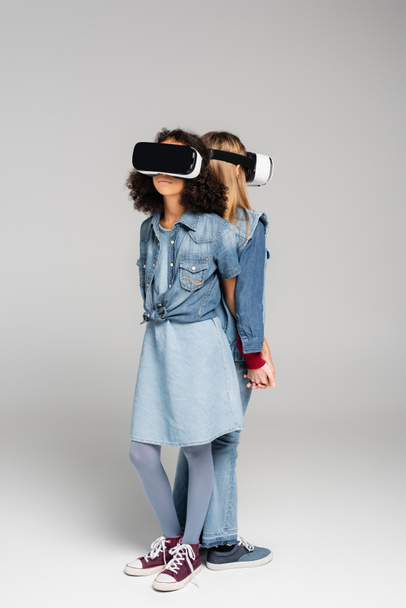 full length view of trendy interracial friends in vr headsets standing back to back on grey - Photo, Image