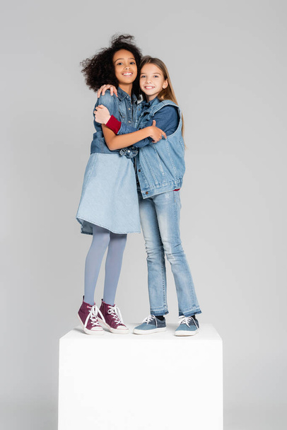joyful multicultural girls in trendy denim clothes embracing on white cube isolated on grey - Foto, Bild