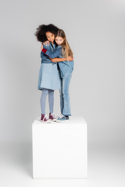 astonished interracial girls in trendy denim clothes embracing while standing on white cube on grey - Fotó, kép