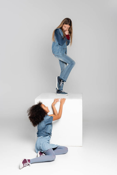 cheerful girl standing on white cube while african american girl sitting on grey - Photo, Image