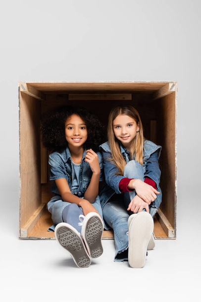 μοντέρνα διαφυλετικά κορίτσια χαμογελώντας στην κάμερα, ενώ κάθεται μέσα σε κουτί σε γκρι - Φωτογραφία, εικόνα