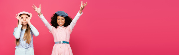 excited interracial girls in trendy clothes and caps posing isolated on pink, banner - Fotó, kép