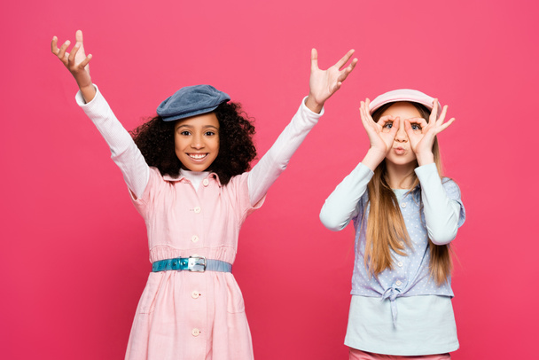 trendy girl imitating eyeglasses near cheerful african american girl with raised hands isolated on pink - Valokuva, kuva
