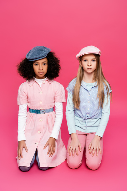 multicultural girls in fashionable clothes and caps looking at camera while posing on pink - Фото, изображение