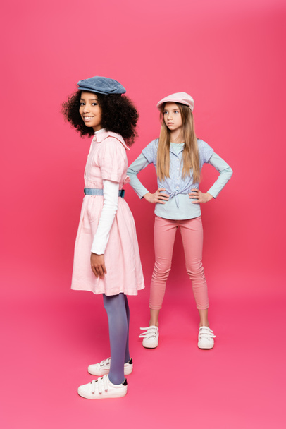stylish girl posing with hands on hips near happy african american girl looking at camera on pink - Photo, Image