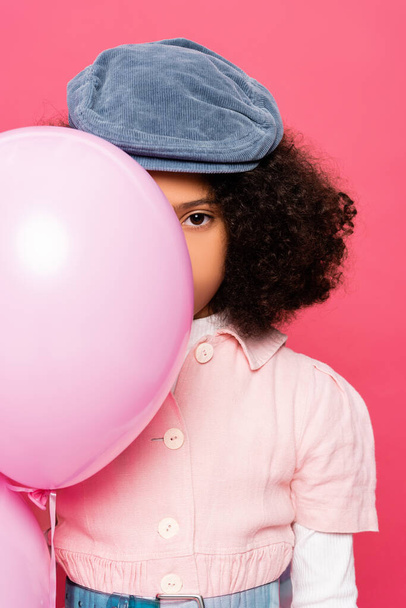 modieus Afrikaans amerikaans meisje in stijlvolle cap obscuring gezicht met feestelijke ballon geïsoleerd op roze - Foto, afbeelding