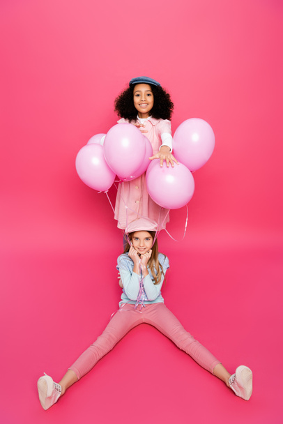 menina americana africana feliz perto de amigo na moda sentado com balões festivos isolados em rosa  - Foto, Imagem