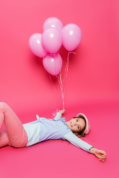 ピンクのお祝いの風船の近くに横たわっている間にカメラで微笑む流行の服の明るい子供 - 写真・画像