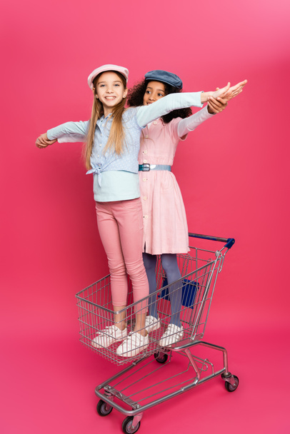 stylish multicultural kids having fun in shopping cart on pink - Foto, imagen