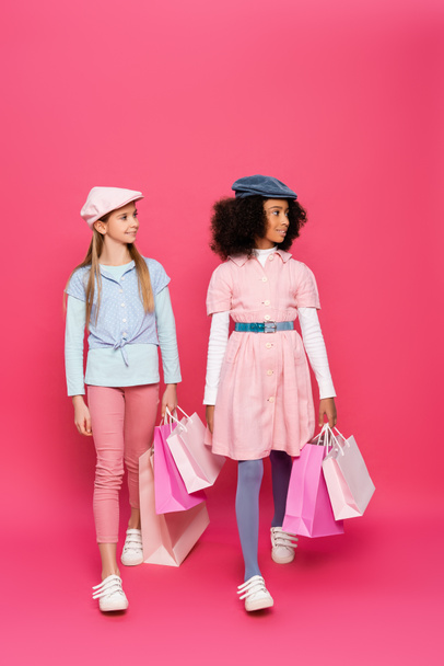 fashionable interracial girls walking with shopping bags on pink - Zdjęcie, obraz