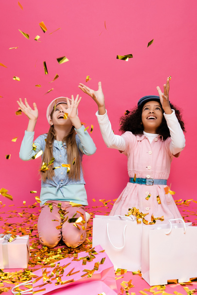 изумленные, модные межрасовые девушки бросают конфетти рядом с подарками на розовый - Фото, изображение