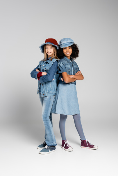 stylish interracial girls in denim clothes and gumshoes standing with crossed arms on grey - Fotó, kép