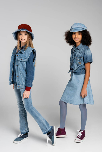 two interracial girls in fashionable denim clothes and hats posing on grey - Foto, afbeelding