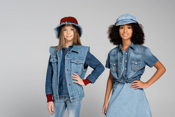 fashionable interracial kids in denim clothes and hats posing with hands on hips on grey - Fotó, kép
