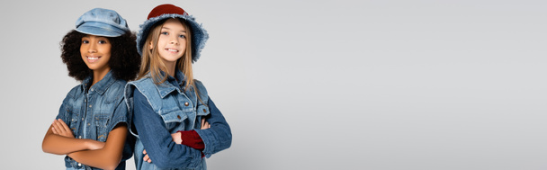 счастливые межрасовые девушки в джинсовой одежде и шляпах позируют со скрещенными руками изолированными на сером, баннер - Фото, изображение
