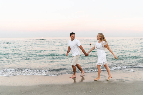 a guy with a girl in summer clothes are walking by the ocean. husband and wife at a romantic sunset of the day near the sea. lovers run along the beach. honeymoon for the newlyweds. - Фото, зображення