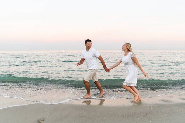 a guy with a girl in summer clothes are walking by the ocean. husband and wife at a romantic sunset of the day near the sea. lovers run along the beach. honeymoon for the newlyweds. - Foto, Imagem