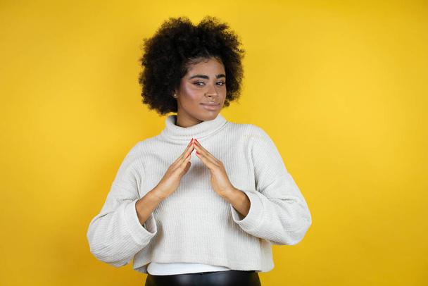Afro-Amerikaanse vrouw draagt casual trui over gele achtergrond met handen bij elkaar en vingers gekruist glimlachend ontspannen en vrolijk. Succes en optimisme - Foto, afbeelding