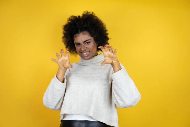 Femme afro-américaine portant un pull décontracté sur fond jaune souriant drôle faisant geste griffe comme chat, expression agressive et sexy - Photo, image