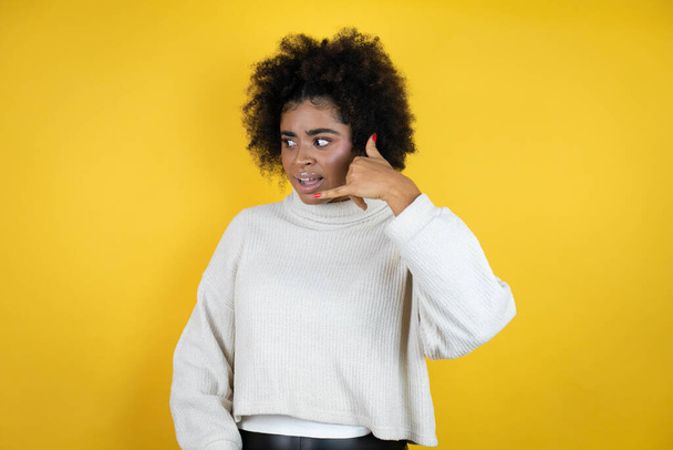 Sarı arka plan üzerine gündelik kazak giyen Afro-Amerikalı kadın telefon hareketlerini telefonla konuşmak gibi elle ve parmaklarla yapıyor. - Fotoğraf, Görsel
