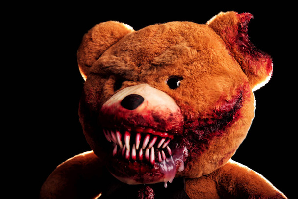 Gruseliger Bär mit einem gruseligen blutigen Grinsen auf schwarzem Hintergrund. Horrorspielzeug. Halloween-Konzept. eine abgetrennte Pfote und ein abgebissenes, blutüberströmtes Ohr - Foto, Bild