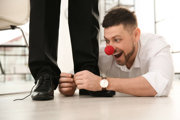 Man with clown nose tying shoe laces of his colleague together in office, closeup. Funny joke - 写真・画像