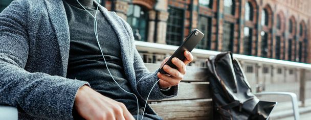 Modern businessman with smartphone in his hands on a city street. Handsome man wearing earphones and speaking on mobile phone, listening music or podcast outdoors. Close up - Photo, Image